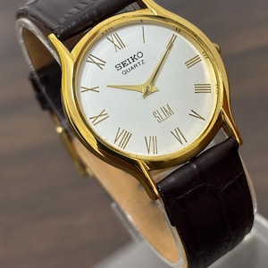May include: A gold-toned Seiko Quartz Slim watch with a white face and Roman numeral hour markers. The watch has gold hands and a dark brown leather band. The watch is displayed on a clear stand.