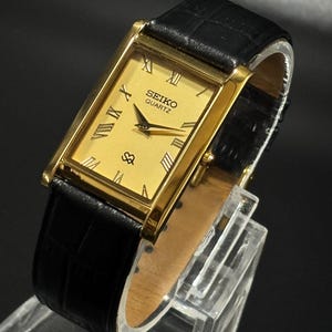 Könnte beinhalten: Eine goldfarbene Seiko Quartz-Uhr mit rechteckigem Zifferblatt und schwarzem Lederarmband. Das Zifferblatt der Uhr zeigt römische Ziffern und ein kleines Sekunden-Zifferblatt. Die Uhr wird auf einem transparenten Ständer präsentiert.