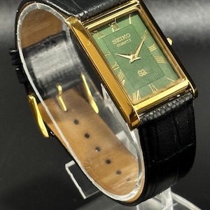 May include: A gold-toned rectangular Seiko Quartz watch with a green face and Roman numerals. The watch has a black leather band. The watch face has a textured pattern and the Seiko Quartz logo.