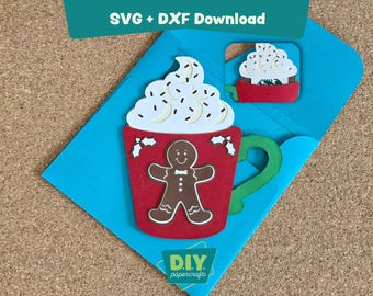 May include: A festive DIY papercraft card featuring a red mug with a gingerbread man, whipped cream, and holly accents. The card is on a blue background with the text "SVG + DXF Download" and "DIY papercrafts".