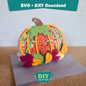 Peut inclure: Une citrouille en papier découpé avec des accents orange, verts et marron, remplie de paillettes. La citrouille est décorée de feuilles vertes et de feuillage d'automne coloré. Le texte "SVG + DXF Download" et "DIY papercrafts" est inclus.