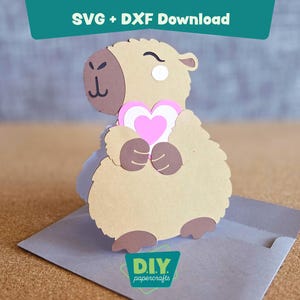 May include: A papercraft capybara holding a pink and white heart. The capybara is light brown with darker brown accents and is sitting on a gray envelope. The image includes the text "SVG + DXF Download" and "DIY papercrafts".