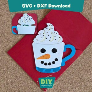 May include: A white papercraft of a snowman in a mug with a blue handle and a carrot nose, set on a red envelope. The mug is topped with whipped cream and sprinkles. The text "SVG + DXF Download" is at the top.