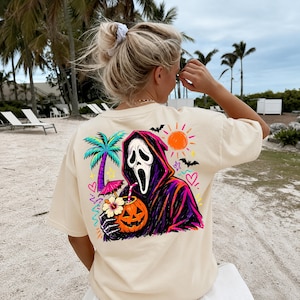 Pode incluir: Uma t-shirt de manga curta cor de creme com um gr&aacute;fico nas costas de uma figura encapuzada com m&aacute;scara branca que segura uma bebida de ab&oacute;bora com palhinha e chap&eacute;u-de-sol, incluindo uma palmeira, um sol laranja brilhante e morcegos.