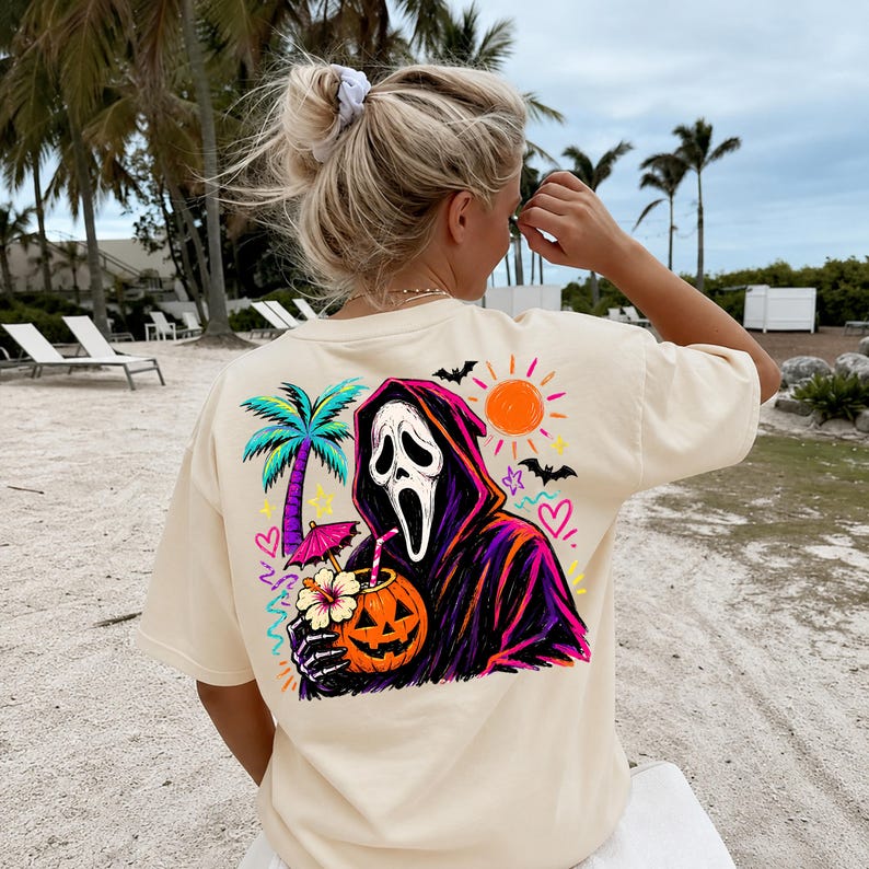 Pode incluir: Uma t-shirt de manga curta cor de creme com um gr&aacute;fico nas costas de uma figura encapuzada com m&aacute;scara branca que segura uma bebida de ab&oacute;bora com palhinha e chap&eacute;u-de-sol, incluindo uma palmeira, um sol laranja brilhante e morcegos.