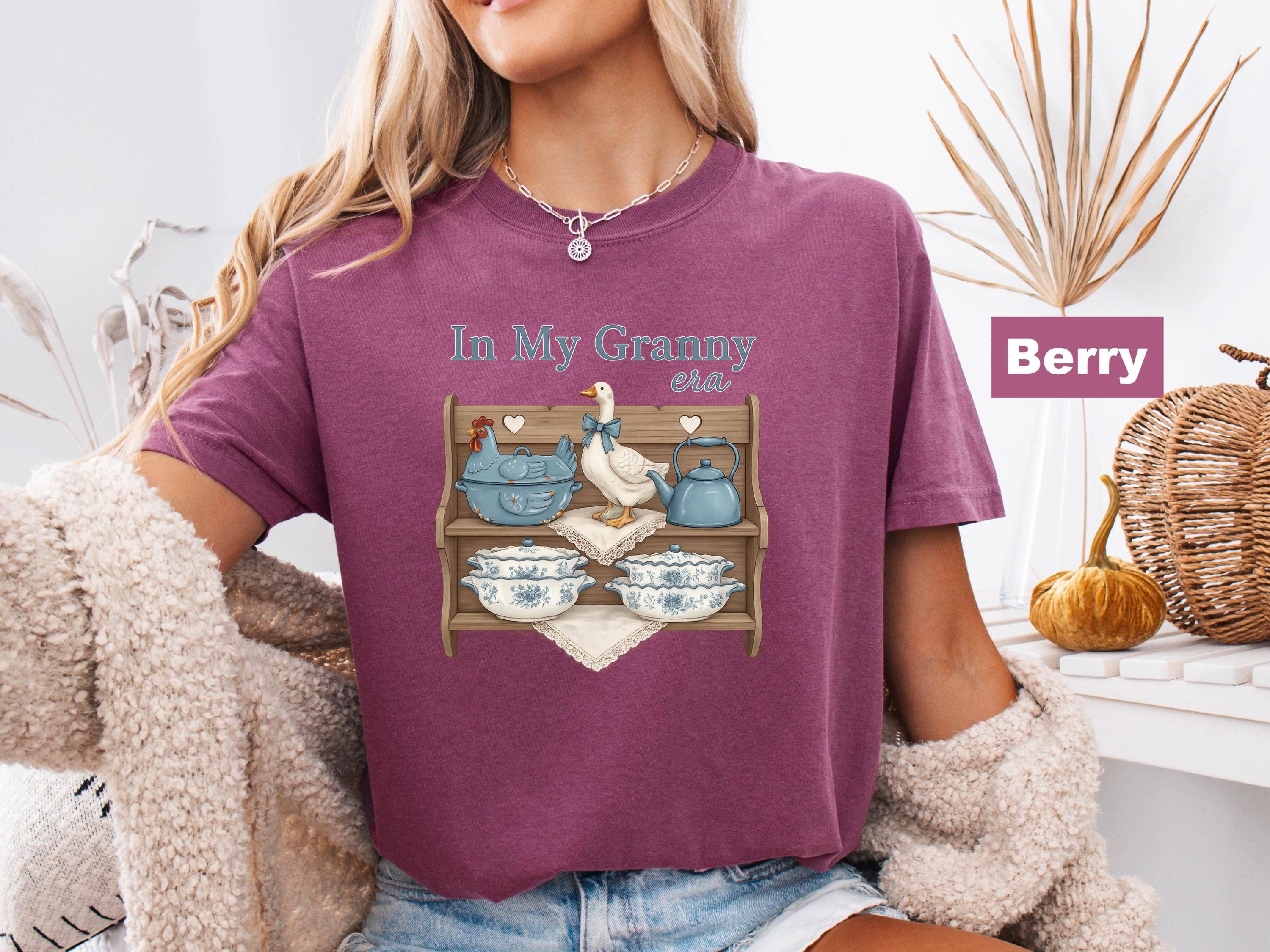 May include: Berry-colored t-shirt with the text "In My Granny era." The shirt features a graphic of a wooden shelf with a blue chicken, a white duck, a blue teapot, and floral-patterned bowls.