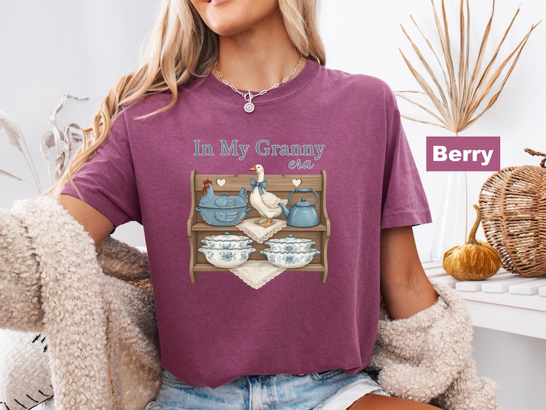 May include: Berry-colored t-shirt with the text "In My Granny era." The shirt features a graphic of a wooden shelf with a blue chicken, a white duck, a blue teapot, and floral-patterned bowls.