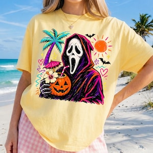 Pode incluir: Uma t-shirt de manga curta amarelo-p&aacute;lido com uma impress&atilde;o gr&aacute;fica de uma figura encapuzada com m&aacute;scara, segurando uma bebida de ab&oacute;bora com um guarda-chuva tropical e palhinha, rodeada por uma palmeira, um sol, morcegos e cora&ccedil;&otilde;es coloridos.
