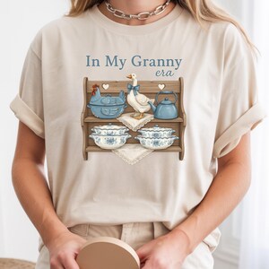 May include: Beige t-shirt with the text "In My Granny era" and a graphic of a wooden shelf displaying a blue chicken-shaped dish, a goose, a teapot, and floral bowls. The design has a vintage, farmhouse aesthetic.