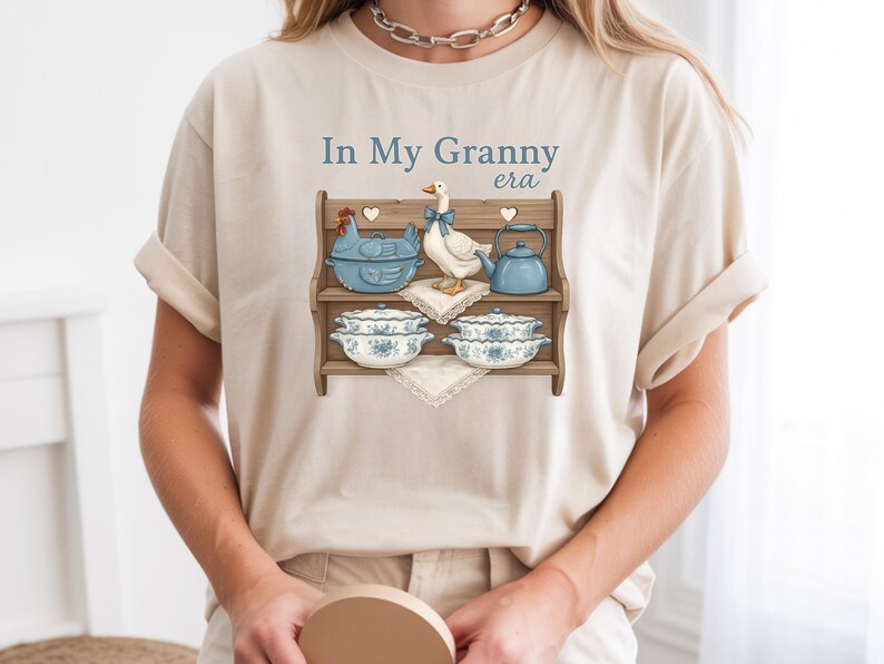 May include: Beige t-shirt with the text "In My Granny era" and a graphic of a wooden shelf displaying a blue chicken-shaped dish, a goose, a teapot, and floral bowls. The design has a vintage, farmhouse aesthetic.