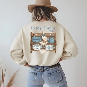 May include: Cream-colored sweatshirt with a graphic of a wooden shelf displaying a blue chicken-shaped dish, a goose, a teapot, and bowls. The text "In My Granny era" is above the shelf. The person is wearing blue jeans and a brown hat.