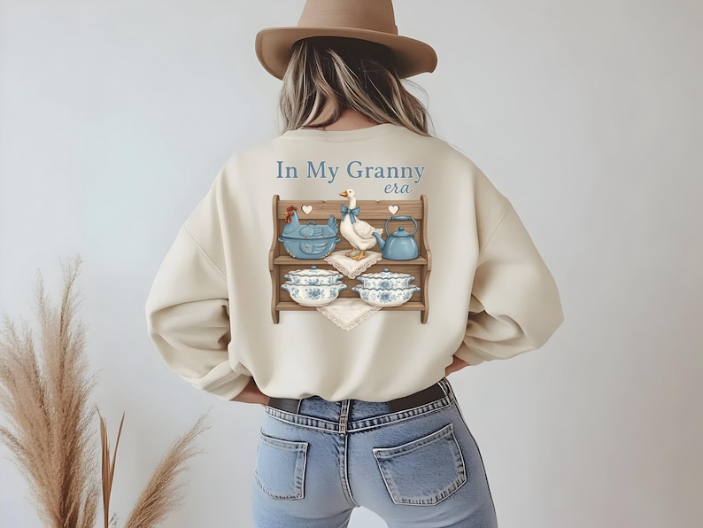 May include: Cream-colored sweatshirt with a graphic of a wooden shelf displaying a blue chicken-shaped dish, a goose, a teapot, and bowls. The text "In My Granny era" is above the shelf. The person is wearing blue jeans and a brown hat.