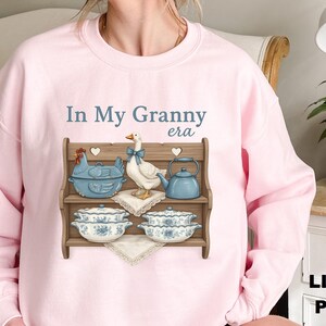May include: Light pink sweatshirt with the text "In My Granny era". The graphic features a wooden shelf with a blue chicken-shaped casserole dish, a goose with a blue bow, a blue teapot, and two decorative bowls.