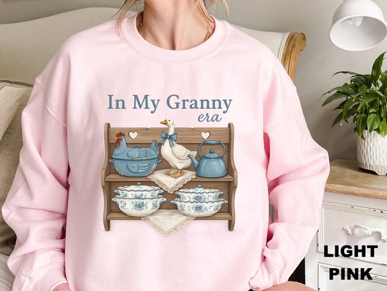 May include: Light pink sweatshirt with the text "In My Granny era". The graphic features a wooden shelf with a blue chicken-shaped casserole dish, a goose with a blue bow, a blue teapot, and two decorative bowls.