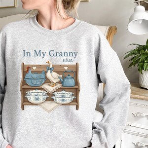 May include: Light gray sweatshirt with the phrase "In My Granny era" in blue text. The graphic features a wooden shelf with a blue chicken-shaped pot, a goose with a blue bow, a blue teapot, and floral-patterned dishes.