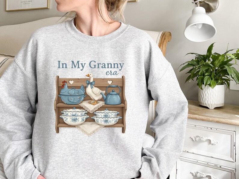 May include: Light gray sweatshirt with the phrase "In My Granny era" in blue text. The graphic features a wooden shelf with a blue chicken-shaped pot, a goose with a blue bow, a blue teapot, and floral-patterned dishes.