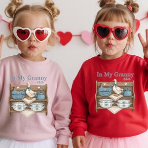 May include: Two children wearing heart-shaped sunglasses and sweatshirts. The sweatshirts are pink and red, each with a graphic of a goose, blue bowls, and the text "In My Granny era". Decorative hearts hang in the background.