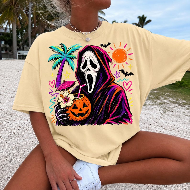 Pode incluir: Uma t-shirt de cor creme com uma estampa gr&aacute;fica de uma figura encapuzada com m&aacute;scara branca segurando uma bebida numa ab&oacute;bora esculpida com palhinha e guarda-chuva. O design inclui uma palmeira, sol, morcegos, cora&ccedil;&otilde;es e estrelas em cores n&eacute;on vibrantes.