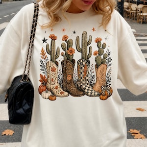 May include: A cream-colored crewneck sweatshirt featuring a graphic print of four cowboy boots, each containing a different cactus with orange flowers. The design includes autumn leaves, small pumpkins, and stars, with a black chain-strap bag on the side.