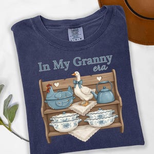 May include: Navy blue t-shirt with the phrase "In My Granny era" in light blue script. The graphic features a wooden shelf with a blue chicken-shaped casserole dish, a goose with a blue bow, and a blue teapot. Two floral dishes are on the bottom shelf.