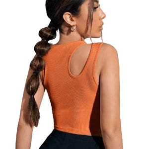 May include: Orange ribbed crop top with a unique shoulder cutout. The sleeveless top is paired with black bottoms. The top is a solid color and has a scoop neckline. The model is wearing gold earrings and has her hair in a ponytail.