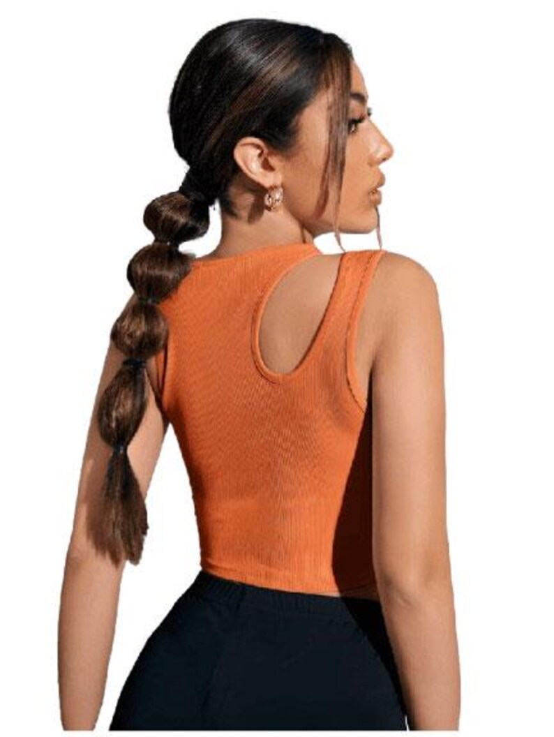 May include: Orange ribbed crop top with a unique shoulder cutout. The sleeveless top is paired with black bottoms. The top is a solid color and has a scoop neckline. The model is wearing gold earrings and has her hair in a ponytail.