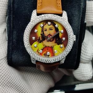 May include: A luxury wristwatch with a brown leather strap and a round face featuring an image of Jesus. The watch has a silver bezel encrusted with small, sparkling stones. The watch is displayed on a black velvet surface.