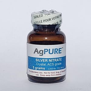 May include: A small brown glass bottle with a white label displaying the text "AgPURE" and "SILVER NITRATE". The label also indicates "5 grams" of the contents. The bottle is sealed with a white cap that has text in English and French.