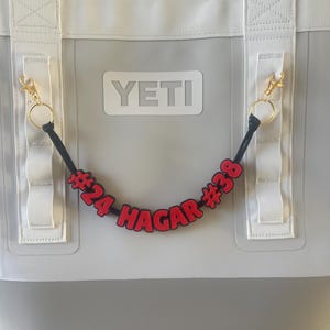 May include: A gray YETI cooler with white straps and gold-colored clips. A black cord with red letters and numbers that read "#24 HAGAR #38" hangs from the cooler. The cooler has a white YETI logo.