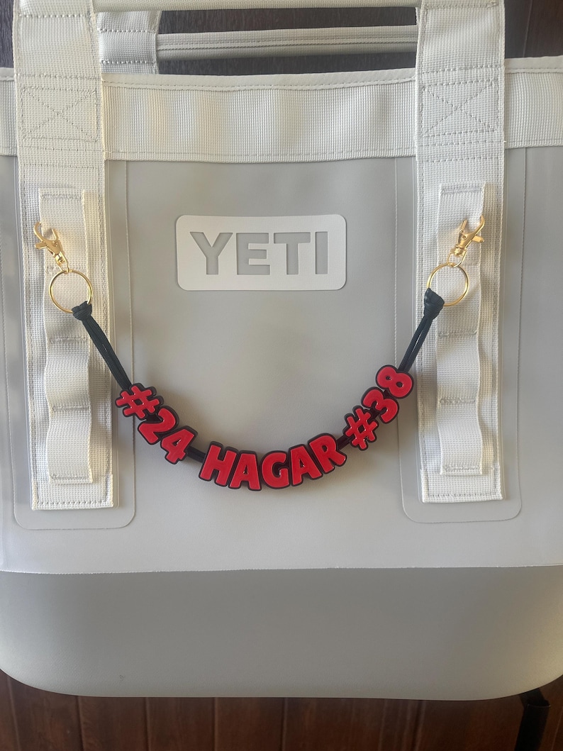 May include: A gray YETI cooler with white straps and gold-colored clips. A black cord with red letters and numbers that read "#24 HAGAR #38" hangs from the cooler. The cooler has a white YETI logo.