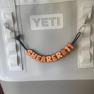 May include: A gray YETI cooler bag with white straps and a black cord with orange letters spelling "SHEARER #11". The letters are attached to a black cord with a metal clasp.