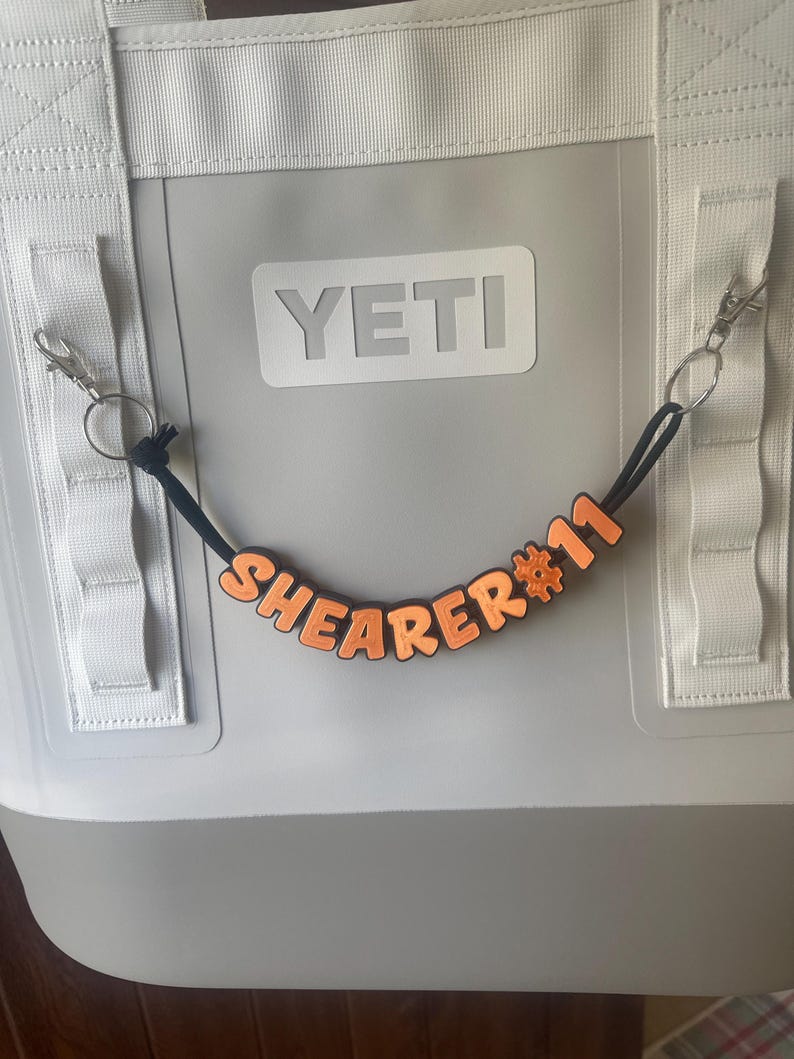 May include: A gray YETI cooler bag with white straps and a black cord with orange letters spelling "SHEARER #11". The letters are attached to a black cord with a metal clasp.