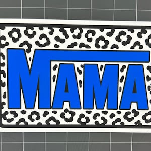 May include: A rectangular sticker with the word "MAMA" in bold blue letters. The letters are outlined in black. The background features a white leopard print pattern with black spots, all within a black border.