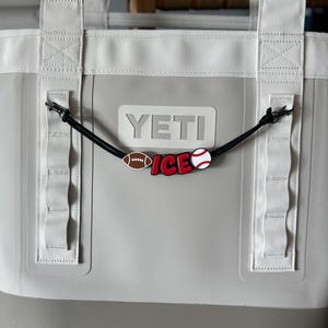 May include: A gray and white YETI tote bag with white handles and a black cord charm. The charm features a football, baseball, and the word "ICE" in red and white.