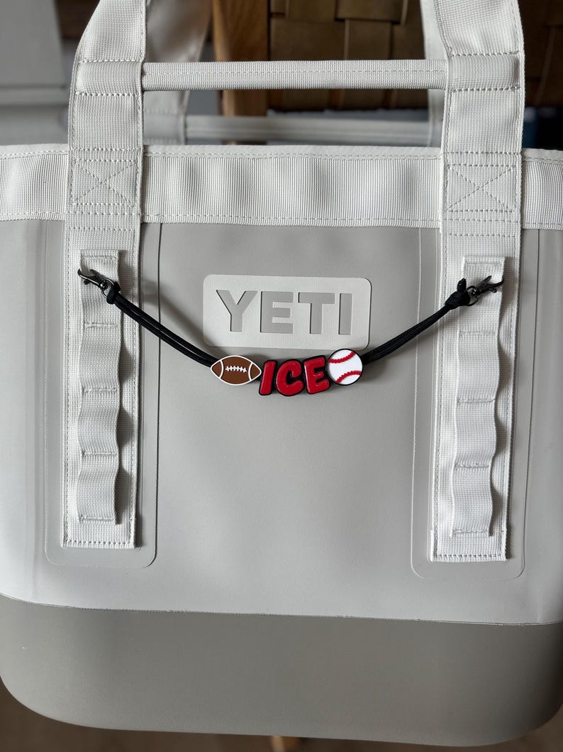 May include: A gray and white YETI tote bag with white handles and a black cord charm. The charm features a football, baseball, and the word "ICE" in red and white.