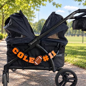 May include: A black double stroller with two adjustable canopies and a black storage pouch attached to the handle. The side of the stroller features orange block letters spelling COLE, a brown football graphic, and the orange number 11.