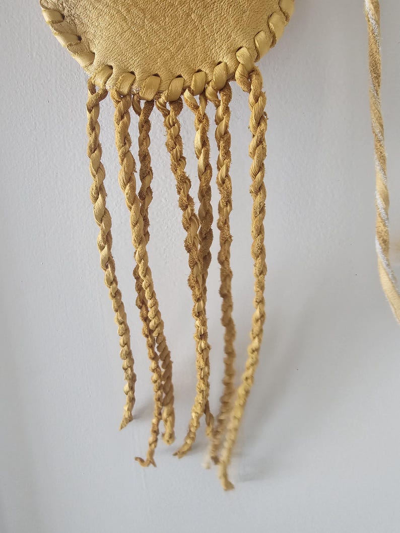 May include: Close-up of a handcrafted, round, light tan leather pouch with braided fringe. The pouch is stitched around the edges and has multiple strands of braided leather hanging from the bottom. The background is a plain white wall.