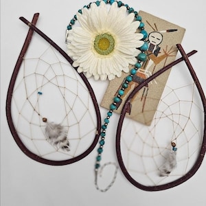 May include: Two teardrop-shaped dreamcatchers made of dark brown twigs with woven tan webbing and grey feathers. A necklace of small turquoise beads and silver spacers rests near a white daisy and a tan card featuring a Kokopelli illustration.