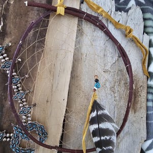 May include: A handcrafted dreamcatcher with a brown circular frame and a woven web. A single black and white feather hangs from the bottom, adorned with beads. A yellow leather strap is attached to the top. A silver necklace with turquoise accents is visible.