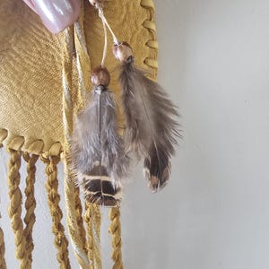 May include: Close-up of a handcrafted, yellow leather bag with braided fringe. The bag features decorative feather and bead accents. A turquoise stone is visible at the top. The bag has a bohemian or Native American aesthetic.