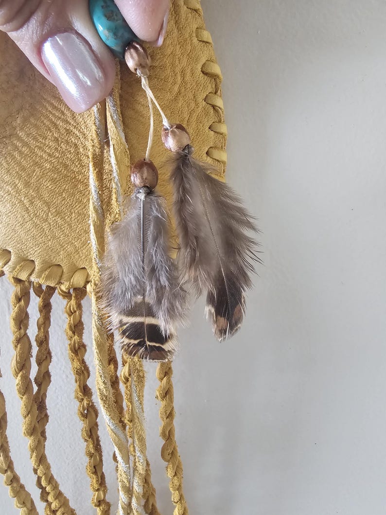 May include: Close-up of a handcrafted, yellow leather bag with braided fringe. The bag features decorative feather and bead accents. A turquoise stone is visible at the top. The bag has a bohemian or Native American aesthetic.