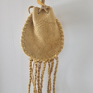 May include: A small, tan leather pouch with a drawstring closure and braided fringe. The pouch is hand-stitched with a decorative stitch around the edges. A twisted cord is attached for hanging. The leather has a natural, textured appearance.