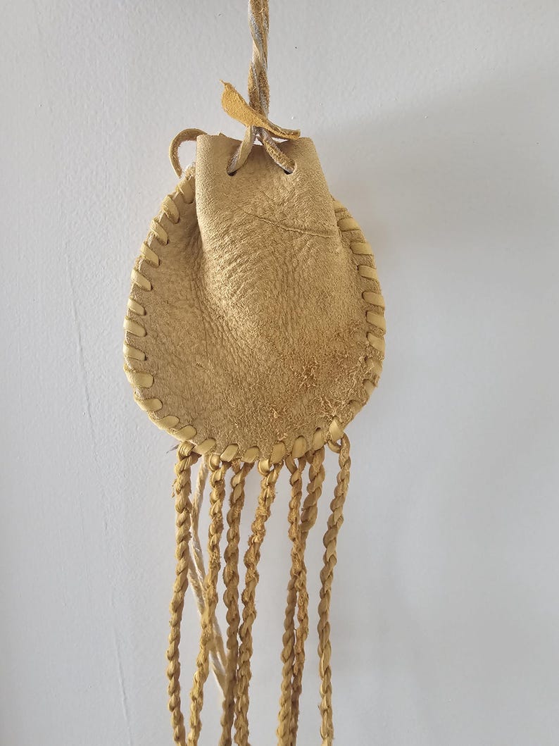 May include: A small, tan leather pouch with a drawstring closure and braided fringe. The pouch is hand-stitched with a decorative stitch around the edges. A twisted cord is attached for hanging. The leather has a natural, textured appearance.
