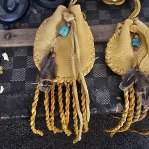 May include: Two small, yellow leather pouches with turquoise beads and feather accents. Each pouch has a braided fringe and a drawstring closure. The pouches are likely handmade and have a rustic, bohemian aesthetic.