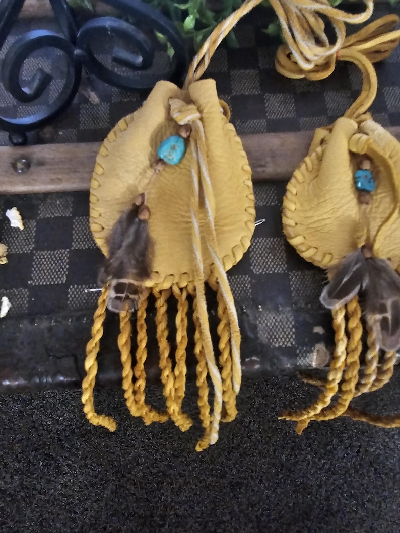 May include: Two small, yellow leather pouches with turquoise beads and feather accents. Each pouch has a braided fringe and a drawstring closure. The pouches are likely handmade and have a rustic, bohemian aesthetic.