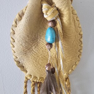 May include: A tan leather pouch with a drawstring closure and braided cord. The pouch is decorated with a turquoise bead, brown beads, and a feather. The edges are stitched with a decorative pattern.