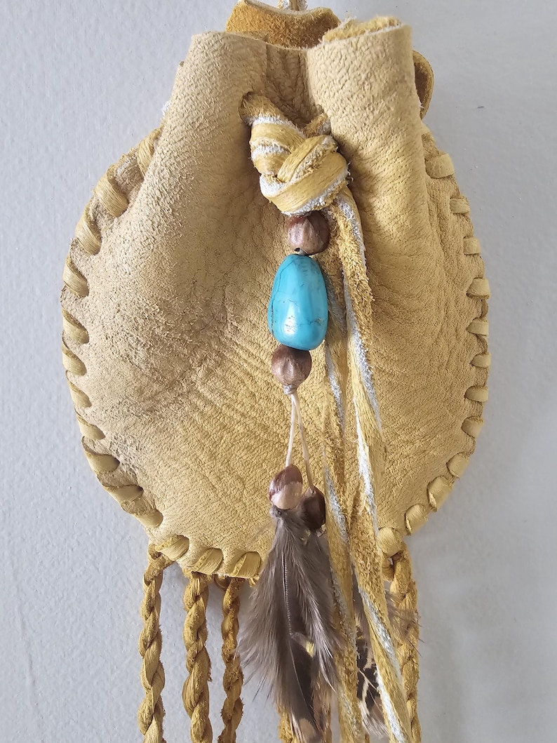 May include: A tan leather pouch with a drawstring closure and braided cord. The pouch is decorated with a turquoise bead, brown beads, and a feather. The edges are stitched with a decorative pattern.