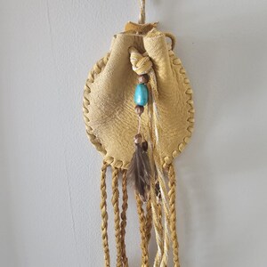 May include: A small, round, tan leather pouch with a drawstring closure. The pouch is decorated with a turquoise bead, brown beads, and a feather. Braided tan and white cords hang from the bottom, and a braided cord is used for hanging.