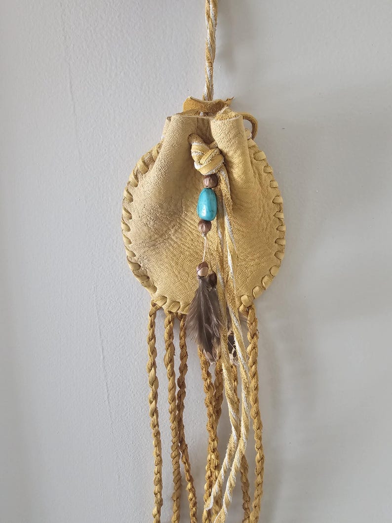 May include: A small, round, tan leather pouch with a drawstring closure. The pouch is decorated with a turquoise bead, brown beads, and a feather. Braided tan and white cords hang from the bottom, and a braided cord is used for hanging.