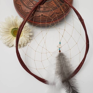 May include: A handcrafted dreamcatcher with a burgundy-colored circular frame and a woven web. It features a cluster of small beads and two feathers, one gray and one white. A decorative terracotta pot and a white daisy are in the background.